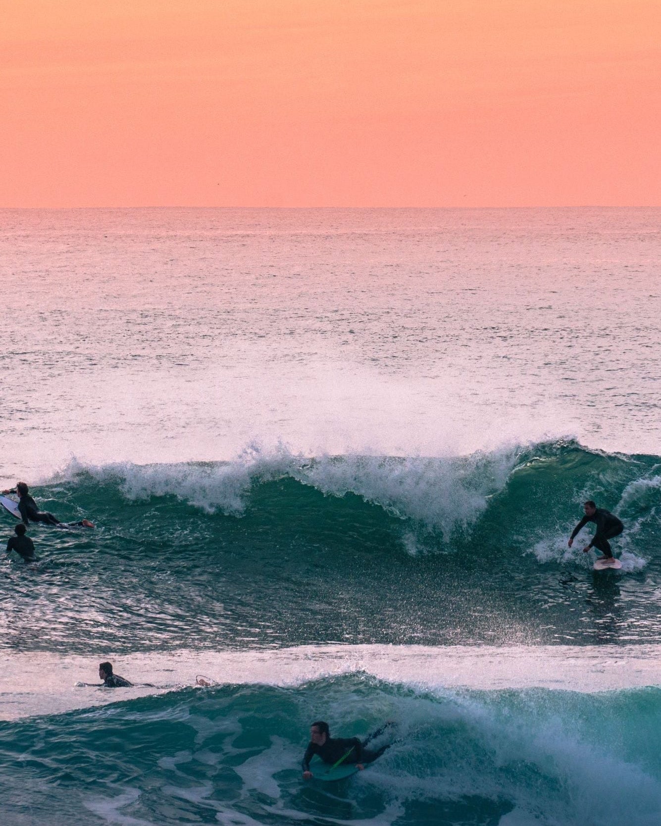 Surfers in the ocean during sunset with a pink and orange sky.